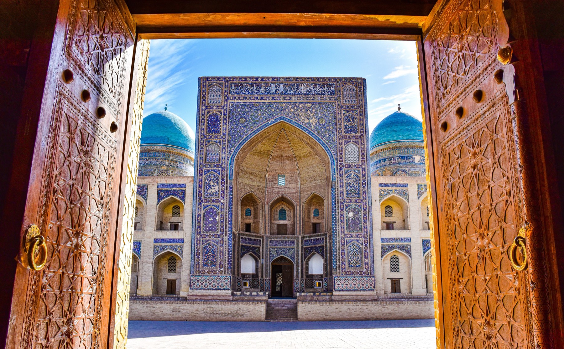 The tiled courtyard of the Kalyan Mosque in Bukhara, framed by a pair of carved wooden doors standing open.