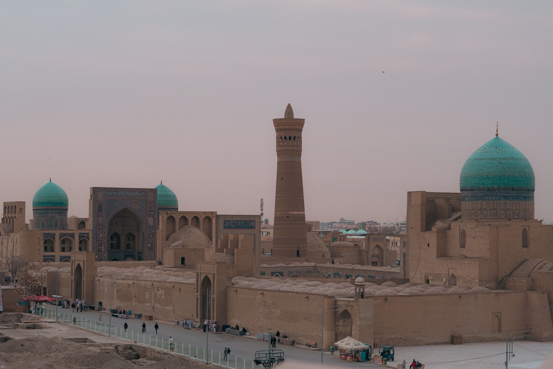 Bukhara's old town skyline at dusk, minaret and turquoise domes above the flat rooftops.