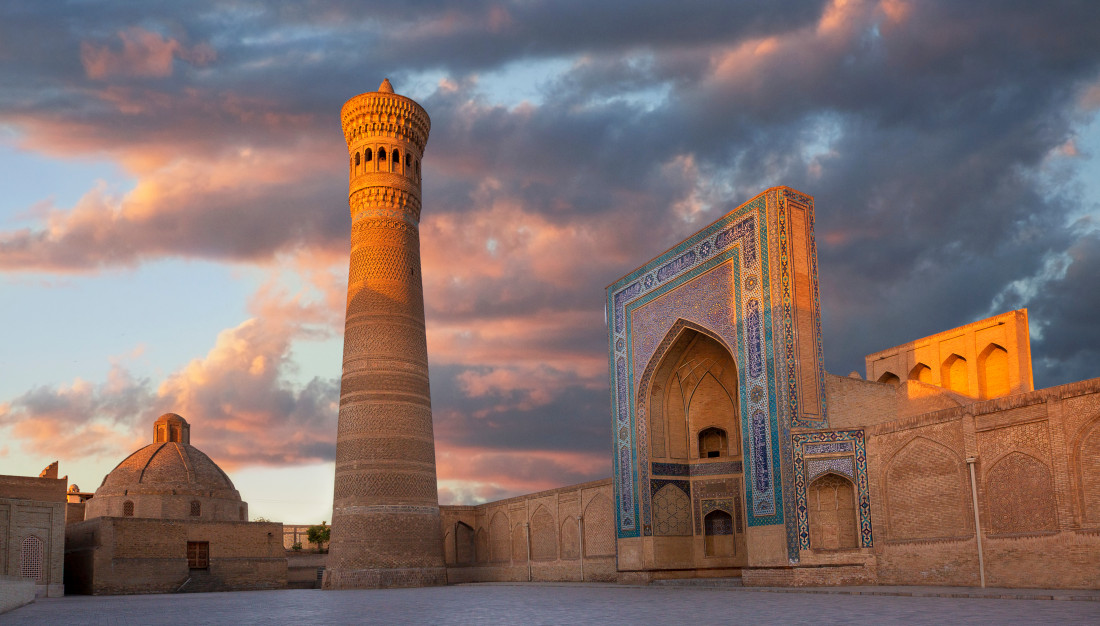 The Kalyan Minaret and the green dome of the Mir-i-Arab Madrasa silhouetted against a burning orange sunset sky over Bukhara.